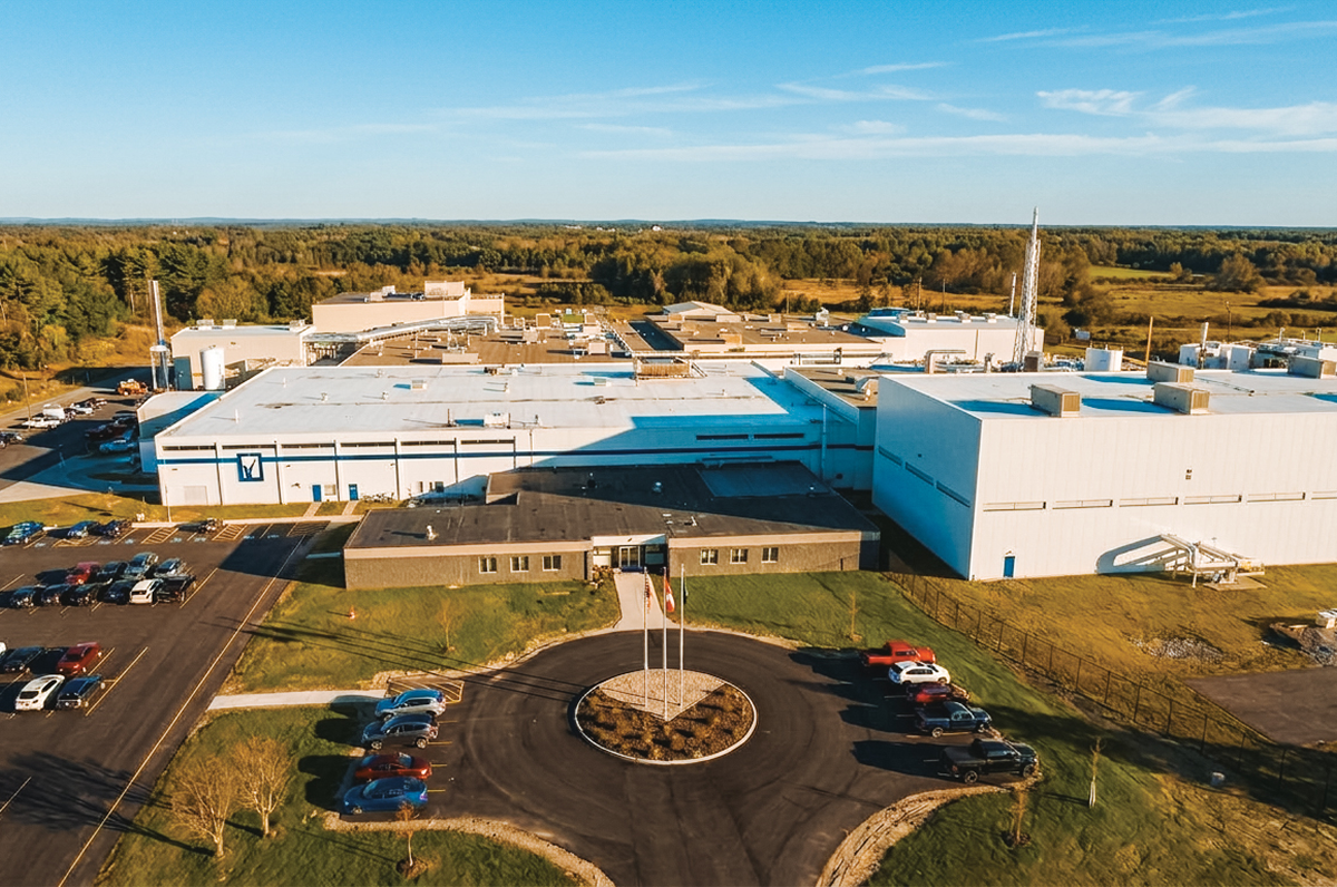 Aerial view of a low white manufacturing facility with a parking lot and circular drive.