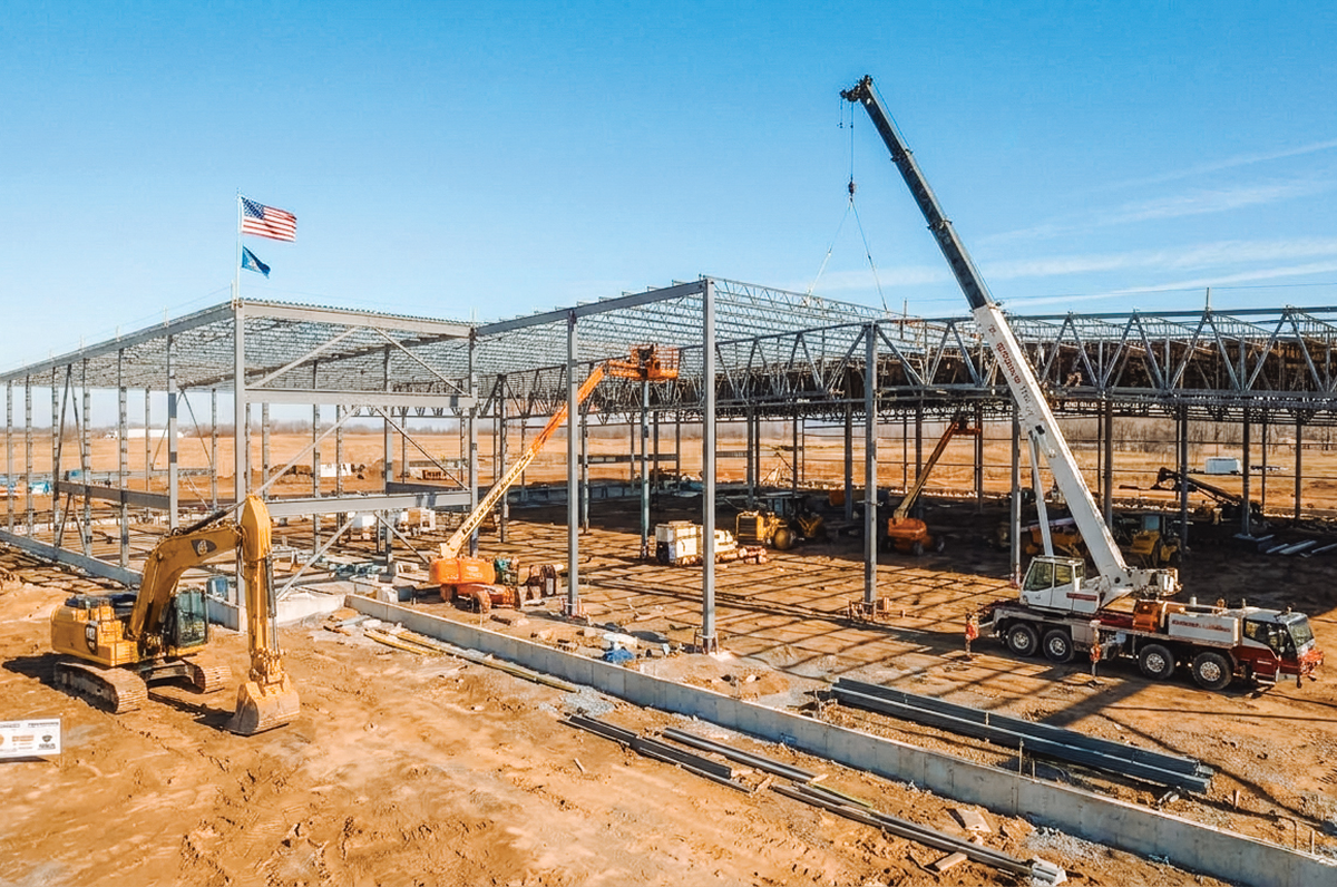 Steel frame of a large building under construction with a crane and excavators under a blue sky.