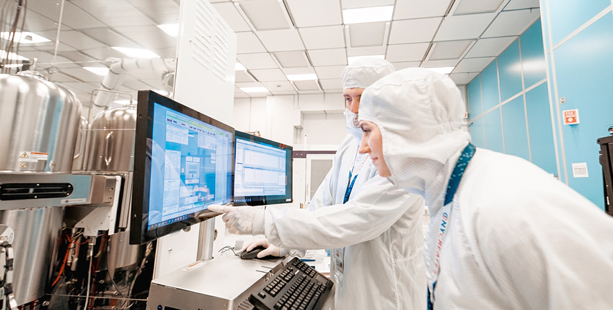 Two researchers in cleanroom suits reviewing data on dual monitors in a lab.