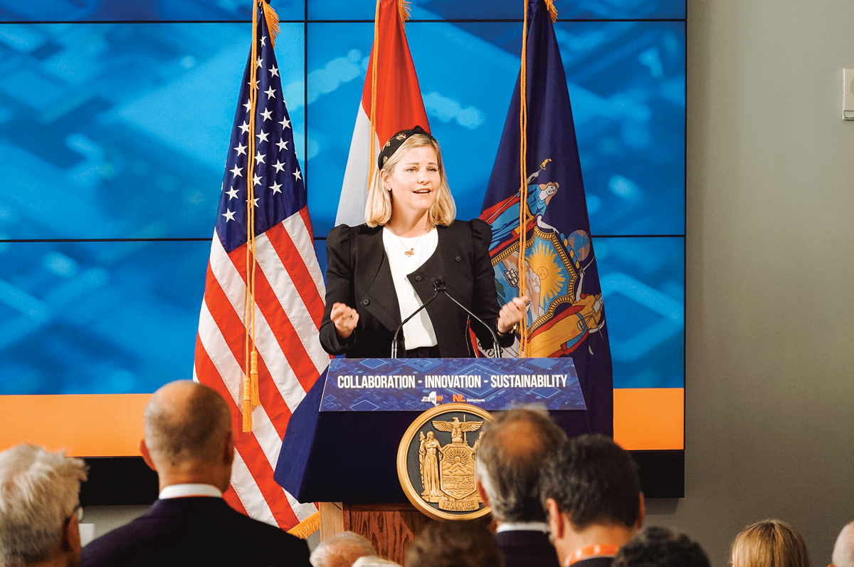 Woman speaking at a podium flanked by U.S., Dutch, and New York State flags.