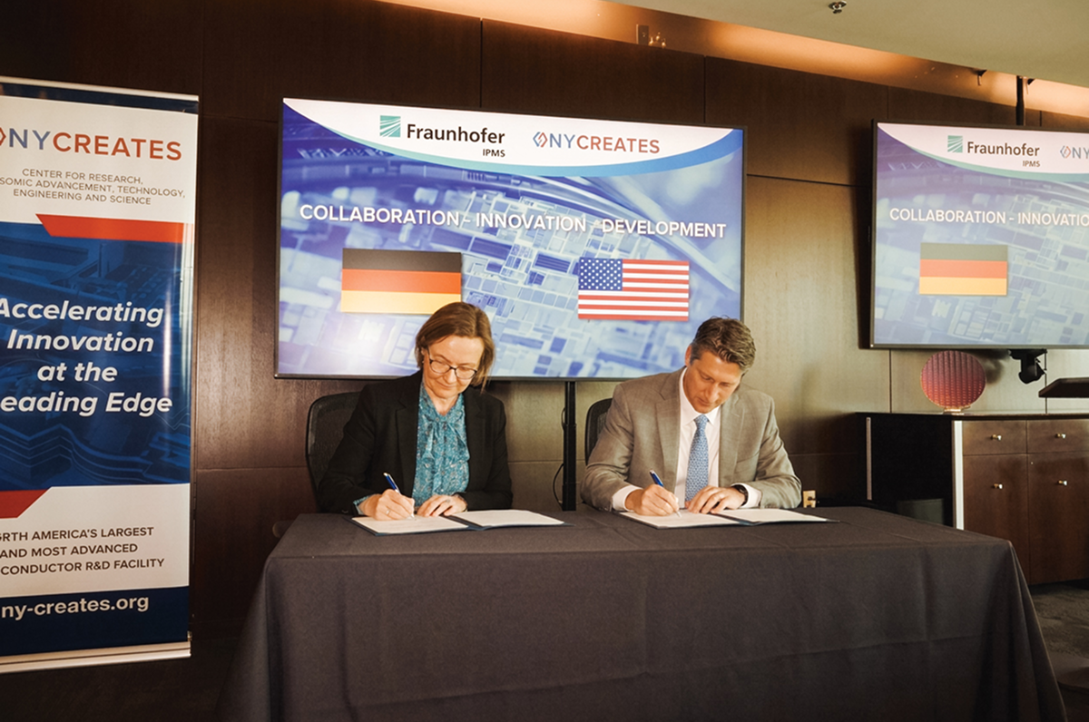 Two people signing documents at a table in front of screens showing Fraunhofer and NY CREATES logos with German and U.S. flags.