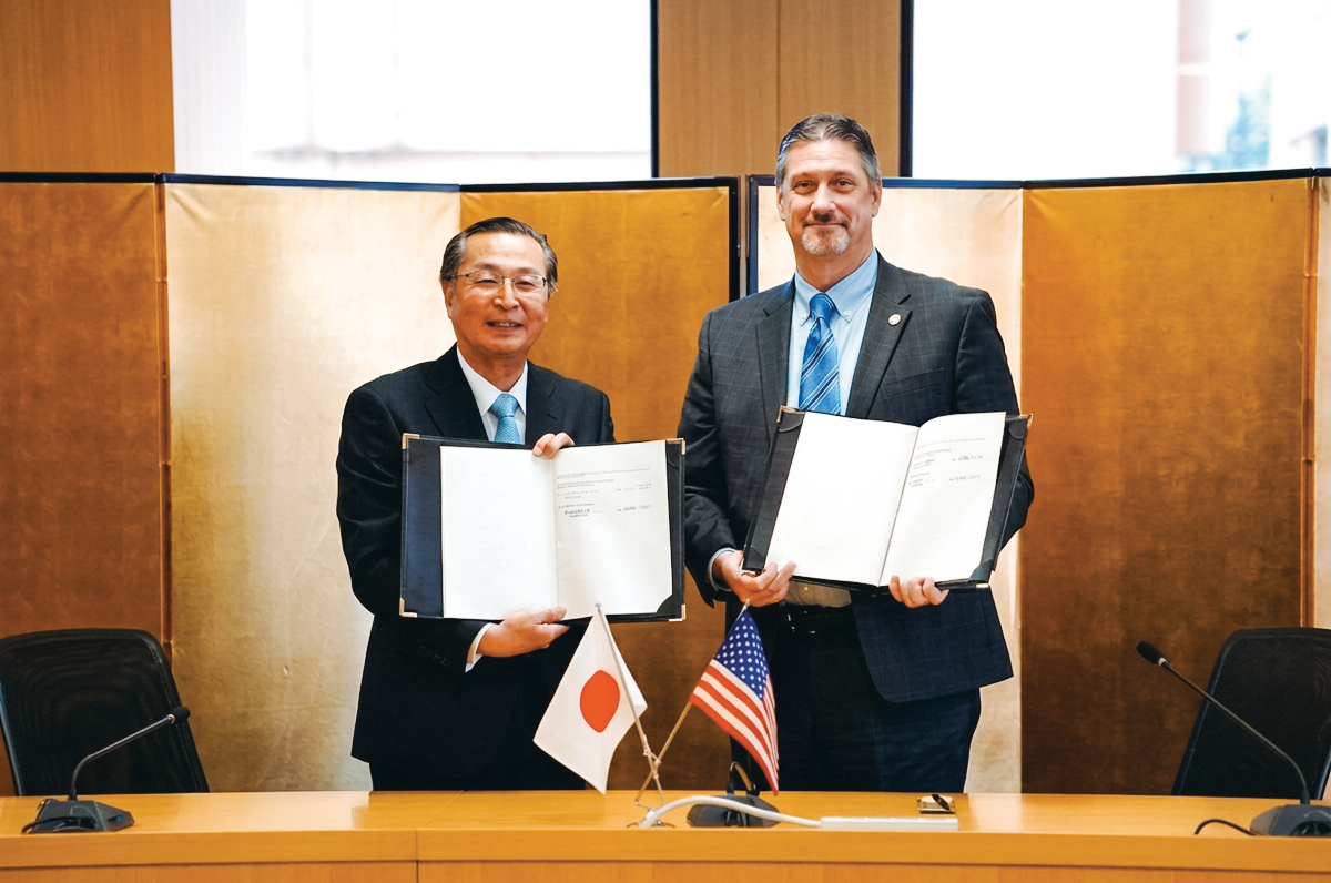 Two men holding signed agreements at a table with small Japanese and U.S. flags.